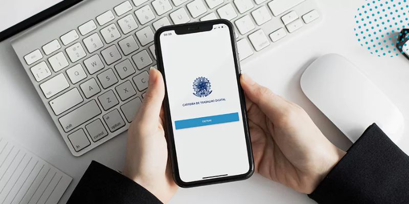 Duas mãos seguram um celular sobre um teclado de computador, destacando a tela da Carteira de Trabalho Digital, simbolizando a modernização e a facilidade de acesso aos serviços do MTE de Brazlândia.