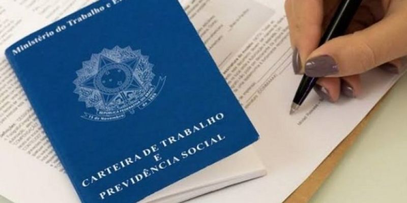 Uma Carteira de Trabalho colocada sobre um contrato de trabalho, enquanto uma pessoa assina o contrato, simbolizando o processo de homologação de contratos de trabalho no Ministério do Trabalho de Brazlândia.